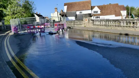 A pool of water covering a bridge with metal fencing blocking the way through for cars. It is a sunny day with blue skies.