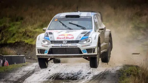Geograph / Brian Deegan A white VW rally car with strips of blue. The Red Bull logo is written on the front. We can see rugged grass land around. Mud is shooting into the air. It's a dramatic image of the car flying over a hill. 