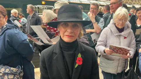 Wera Hobhouse, MP for Bath, dressed in a black hat, black turtleneck and a black blazer with a poppy on the lapel. She stands on a railway platform. There are lots of people in the background.