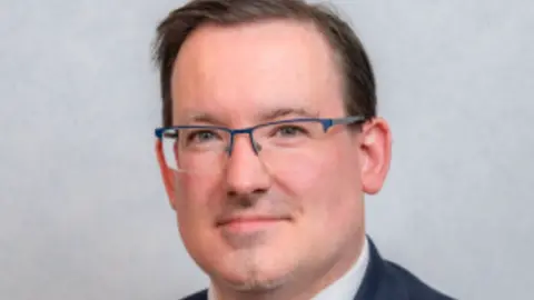 Tynwald Headshot of Lawrie Hooper - he wears glasses, has short hair and is smiling to the camera.