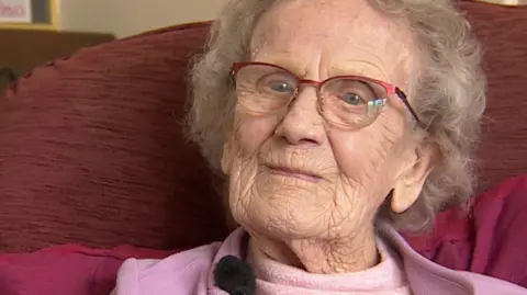 Dorothy Howard is sitting in a chair and wearing a pink fleece and t-shirt. She has medium length white hair and pink-trimmed glasses.