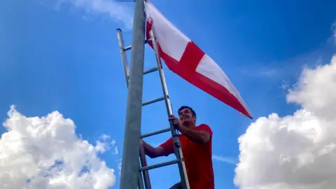 BBC/Mousumi Bakshi A man in a red T-shirt mounts a ladder to fix a St George's flag to a lamppost.