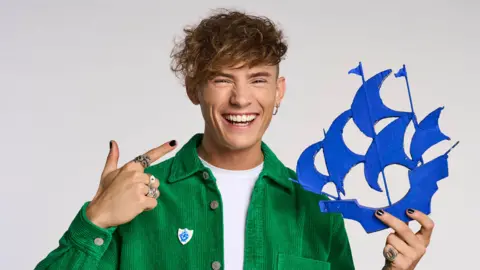 A man with dark curly hair is smiling and looking into the camera. He's dressed in a green shirt, white t-shirt and multiple rings. He's holding a cut out of the blue Blue Peter ship and point to it.