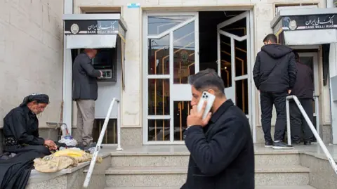 Men using ATMS and another man on his phone, outside a bank.