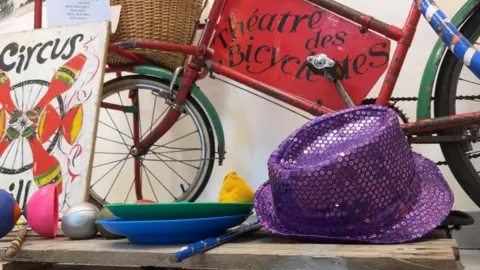 Items arranged on a table for an exhibition - a purple sequinned hat, old painted bicycle and equipment for circus tricks, like spinning plates