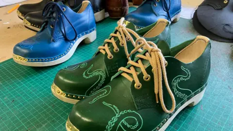 Katie Galbraith A pair of green clogs placed on a workbench. They are decorated with neat swirls.