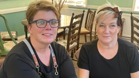 Karen Wattam and Anne Ward sitting inside their cafe. They are both wearing black T-shirts. Karen has short hair and blue rimmed glasses, while Anne has short blonde hair with a brown bow. There are chairs and tables in the background.