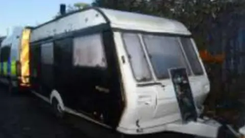 West Midlands Police A caravan with black sides and a white roof and front, is parked along a fence, with a police van parked directly behind it.