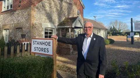 Clare Worden/BBC Terry Stork is wearing a black suit and white shirt. He is leaning on a sign which says "Stanhoe Stores" in front of a building which is clad with lots of round stones
