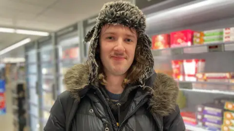Simon Thake A young man with a light moustache and wearing a deer stalker hat stands in-front of a supermarket freezer door
