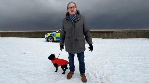 Kevin Shoesmith/BBC A man in his 70s, wearing a hat, scarf, gloves and a thick coat, stands on a promenade under a snowy sky. Snow is on the ground.