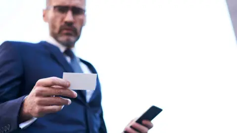 A man, in a blurred image, wearing a dark blue suit, holding a mobile phone and bank card. It is a stock image 