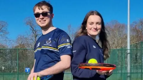 Dogs Trust Ruby Pummell and Ed Ingles wearing blue tops and holding tennis rackets and balls. Ed has brown hair and is wearing sunglasses. Ruby has long brown hair. Both are smiling