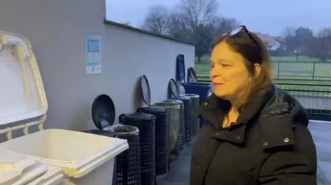 A woman with glasses on her head wearing a black hooded quilted coat standing next to a white bin, with seven other bins visible next to it