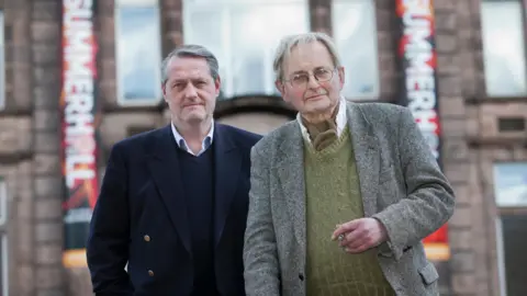 Alamy Iain Gale, a grey-haired man with a strong jaw, is on the left wearing a blue, gold-buttoned, double-breasted blazer, light blue open-necked shirt and dark blue jumper. To his right, Allan Massie is in a grey, herring-bone tweed jacket, green woollen jumper and white shirt with a brown cravat. Massie is holding a lit cigar. Behind them can be seen a Victorian building with Summerhall branded hangings.
