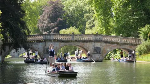 PA Media Four punts filled with people making their way along a river. A stone bridge is in the background. A tree overhangs on the left. A grass bank and shrubs are on the right.