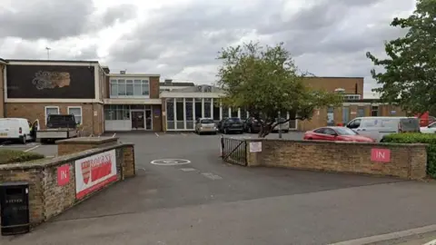 A street view of Bourne Grammar School which shows a brick building with a parking area in front of it. The building has large windows and features a mural of a face on a large dark panel. A wall bordering the building has a banner featuring the word "Bourne" and a red and gold crest. A tree is visible next to a metal gate and the sky is cloudy. 