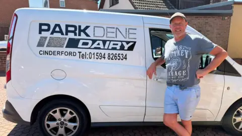 Chris is standing in a housing close beside his white delivery van, which has 'Parkdene Dairy' and a telephone number on the side. He is leaning with an elbow on the driver's window and has his other hand on his hip
