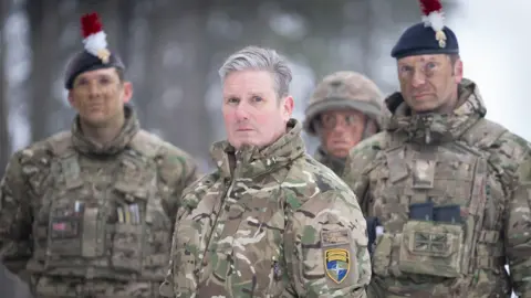 PA Sir Keir Starmer looks at the camera, as do two soldiers standing behind him, during his visit to Estonia. They are all wearing camouflaged jackets.