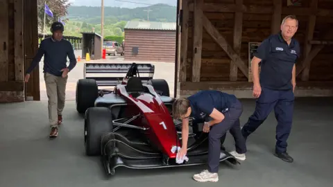 A car being worked on in a shed at the race track. Three men are around the vehicle, one of them is polishing it.
