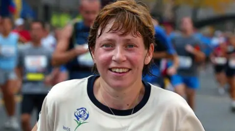 Chris Ison/PA A woman with short brown hair and wearing running gear, looks tired but elated. Behind her are other runners.