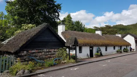Glencoe Folk Museum  Glencoe Folk Museum is low building with white walls and a thatched roof. Behind it are trees. It is a sunny day with white fluffy clouds in the sky.