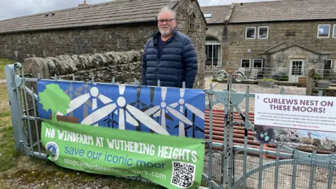 The campaigner stands behind the gateway to his house with a banner tied to the gate. It says No Windfarm At Wuthering Heights.