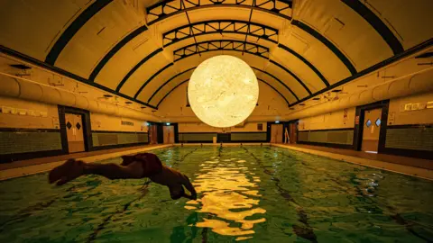 A silhouette of a swimmer diving into an indoor pool. There is a large sculpture modelled off of the sun suspended above the pool. It is reflecting in the water.