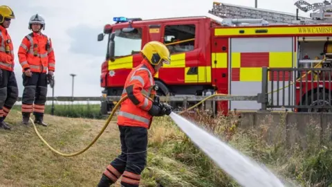 Cambridgeshire Fire and Rescue A firefighter hosing a grassfire with two other firefighters and a fire engine behind him