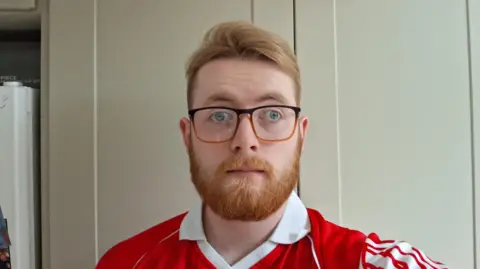 Cathal has ginger hair, lifted at the front, and a well-kept ginger beard. He wears glasses with square frames. His jersey is red with white stripes and a white collar. 