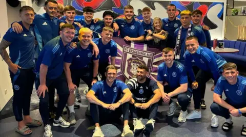 Glenavon FC Glenavon players are arm in arm and smiling. They are wearing navy and blue polo shirts and are holding a purple and white Erzgebirge Aue flag.