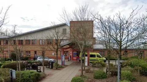 Street view image of Ormskirk Hospital a red brick building with covered walkway through landscaped gardens to the entrance flanked by cars and ambulances