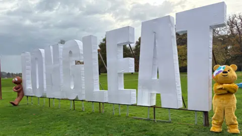 White sign which says 'Longleat' on grass with yellow Pudsey bear stood at one side with his arm crossed and a brown figure, Morph, at the other.