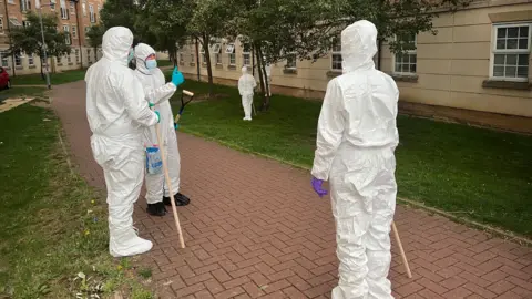 Kate Bradbrook/BBC Three police officers wearing full-body white protective suits stand on a brick pathway bordered by grass. They are near blocks of flats with rows of windows. There are trees beside the path, which runs between the buildings. Two of the officers have poles they have been using to search the area. The third officer has a spade.