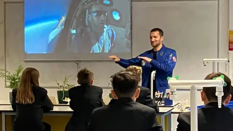 Mr Bennett standing in front of a class of children pointing at images of him in space