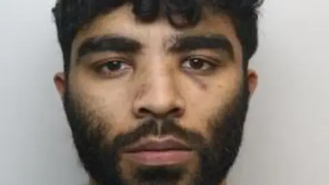 A police mugshot of Hassan Jhangur. He has dark facial hair and dark, slightly curly hair.