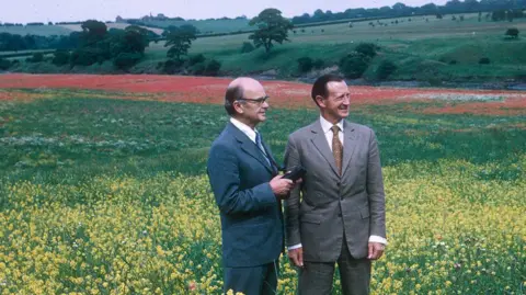 WWT Washington Sir Peter Scott and Sir James Steel observing a colourful meadow at WWT Washington in the 1970s. Sir Peter Scott is wearing a blue suite and tie and white shirt. Sir James Steel is wearing a grey suit with a brown tie and white shirt. The meadow is covered in red, yellow and white flowers.