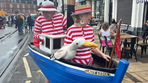 Fleetwood Arts CIC A man and woman in red striped old fashioned bathing suits standing inside a blue boat with a stuffed seagull on the side