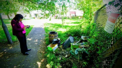 MP Sureena Brackenridge stands next to a pile of rubbish in front of a fence while looking down at her phone.
