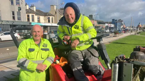 Cathal and Alan are wearing yellow high-vis jackets. They are sitting on red grass cutter machinery and smiling. 

They are both bald and Alan has his navy coat on underneath and grey trousers. His hood is up. 

The town is in the background, shops and the main road are visible. 