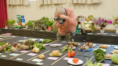National Trust A man with a camera leaning over a table of vegetable entries at a village show