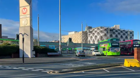 Leeds City Bus Station on a sunny day. There are two buses to the right of the frame - one is green and one is red. The Victoria Gate shopping centre is in the background. 