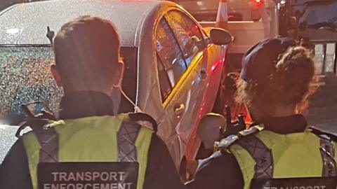 Two transport enforcement officers, one male and one female who is wearing a cap,  stand with their back to camera while looking at a sized grey car being towed away. They are wearing high viz jackets and it is dark