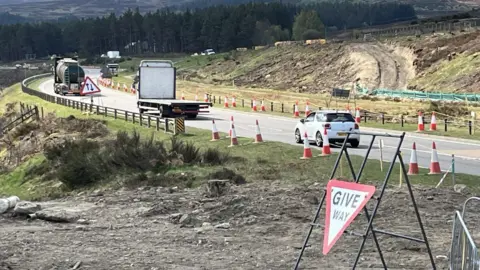 A9 with a queue of moving lorries and cars. There are traffic cones along the side of the road and new tracks can be seen for construction vehicles. There is a give way triangular sign next to the road.  