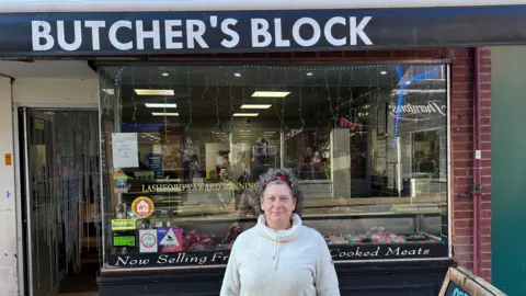 A woman wearing a white jumper stands and looks into the camera. Behind her is a small shop window with glass and the words 'Butcher's Block' written in white letters at the top.