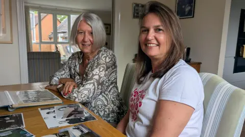 Pat Moody and Sarah Myers look into the camera as they sit at a living room table covered in photos 