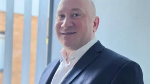 NNUH Man wearing a navy suit jacket and white shirt smiling at the camera. Behind him is a window with blinds.