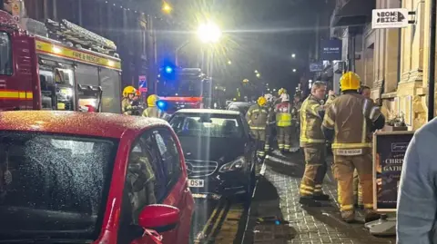 Raja Munuswamy A number of firefighters stand on a street next to parked cars and several fire engines on a street. It is dark and there is a bright street lamp. 