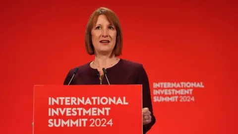 Reuters Rachel Reeves wears a maroon dress as she stands at a red podium which reads: International Investment Summit 