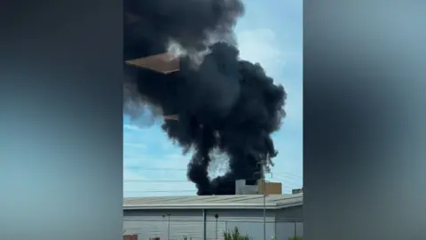 BlueHummingbird2638 A black plume of smoke rising into the air. There is a large industrial unit in the foreground.